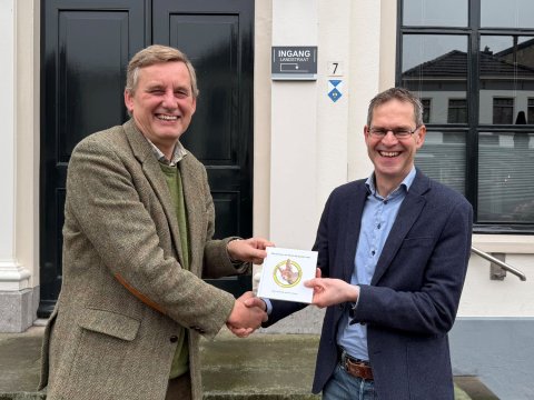 Burgemeester geeft Arjan Klein Nibbelink een hand tijdens overdracht van het voorleesboekje staand op het bordes van het gemeentehuis