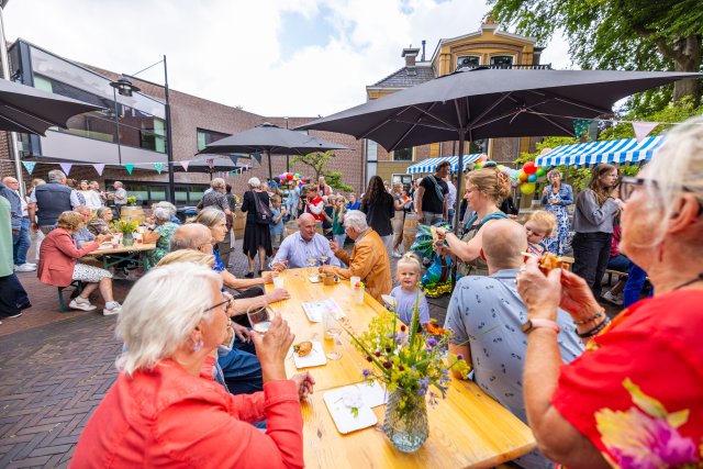 Het plein voor het gemeentehuis vol met tafels met vazen bloemen en bordjes eten, parasols, vlaggetjes en tientallen mensen.