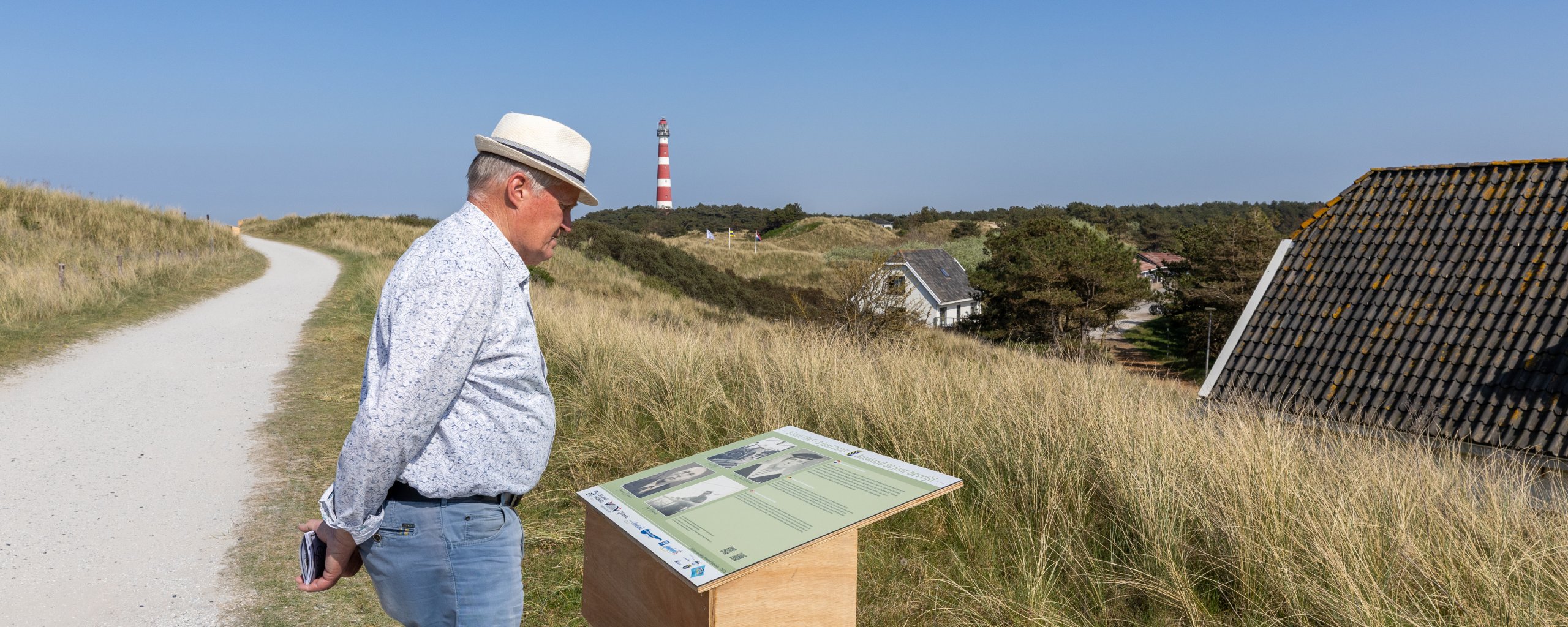 Wethouder Piet IJnsen kijkt bij een herinneringspaneel met op de achtergrond de vuurtoren