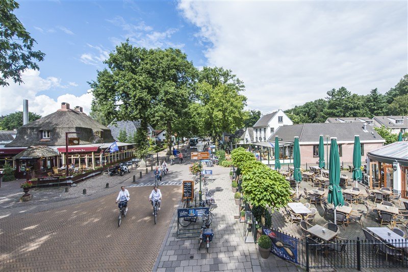 Aanblik van bovenaf op de hoofdstraat van de Lage Vuursche met in het midden twee fietser en aan weerszijden van de weg een aantal horecagelegenheden met bijbehorende terrassen met parasols