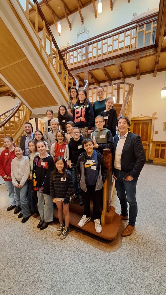 In de hal van de oudbouw van de gemeente Baarn staan 18 kinderen op de trap. Links van de groep wethouder Steven de Vries. Rechts van de groep wethouder Hugo Prakke