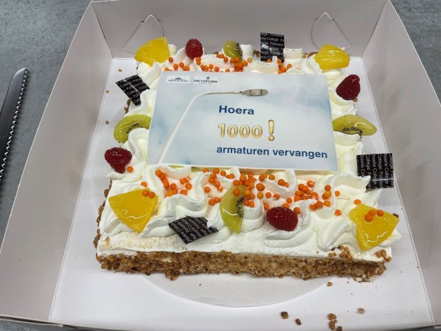 Een taart is van bovenaf gefotografeerd. Op de taart staat de tekst 'Hoera - 1000! armaturen vervangen'.