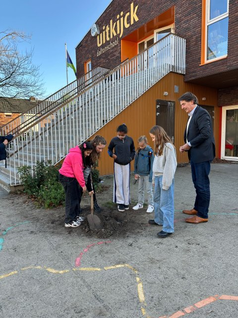 Bij de trap van basisschool De Uitkijck zetten twee leerlingen een schop in de grond en kijken drie andere leerlingen en wethouder Hugo Prakke toe