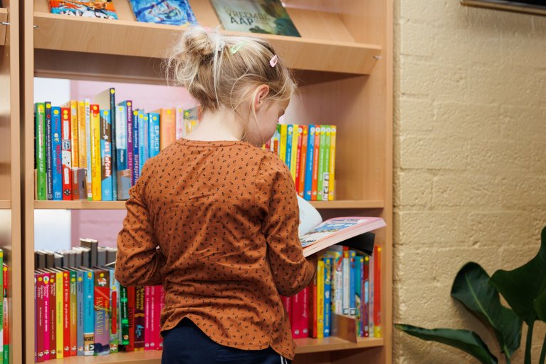 Een meisje staat met haar rug naar je toe in een boek te lezen bij een boekenkast.