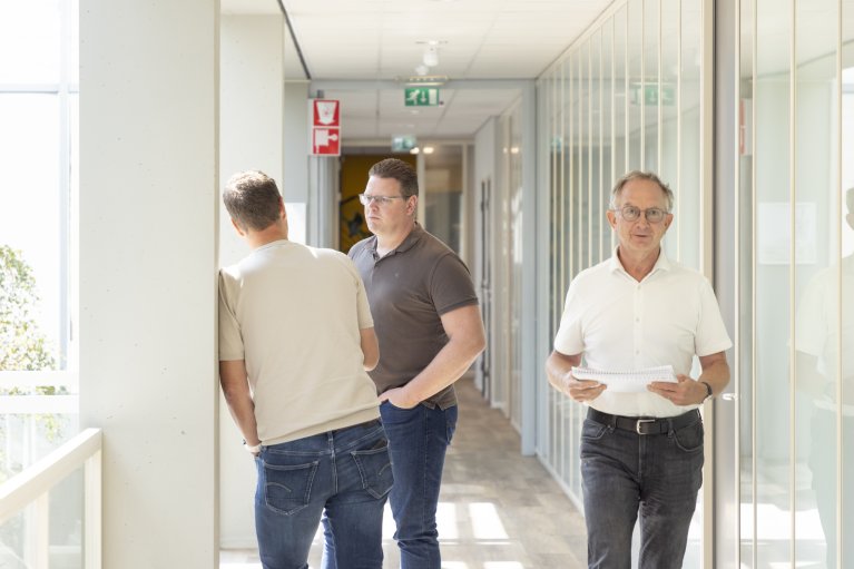 Twee collega's staan te praten op de gang en 1 collega loopt over de gang