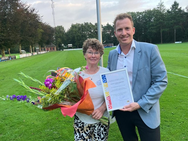 Frieda Verspeek-Moors en wethouder Stef Luijten staan bij elkaar met de onderscheiding en een bos bloemen