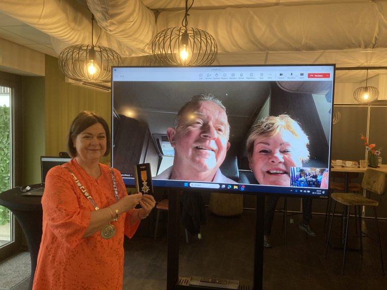 Burgemeester Arinda Callewaert staat met een lintje voor een scherm waarop Hans van den Dungen zichtbaar is