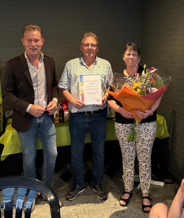 Wethouder Mathijs Kuijken met Geert Wassink met zijn onderscheiding in zijn hand en zijn vrouw met bloemen in de hand