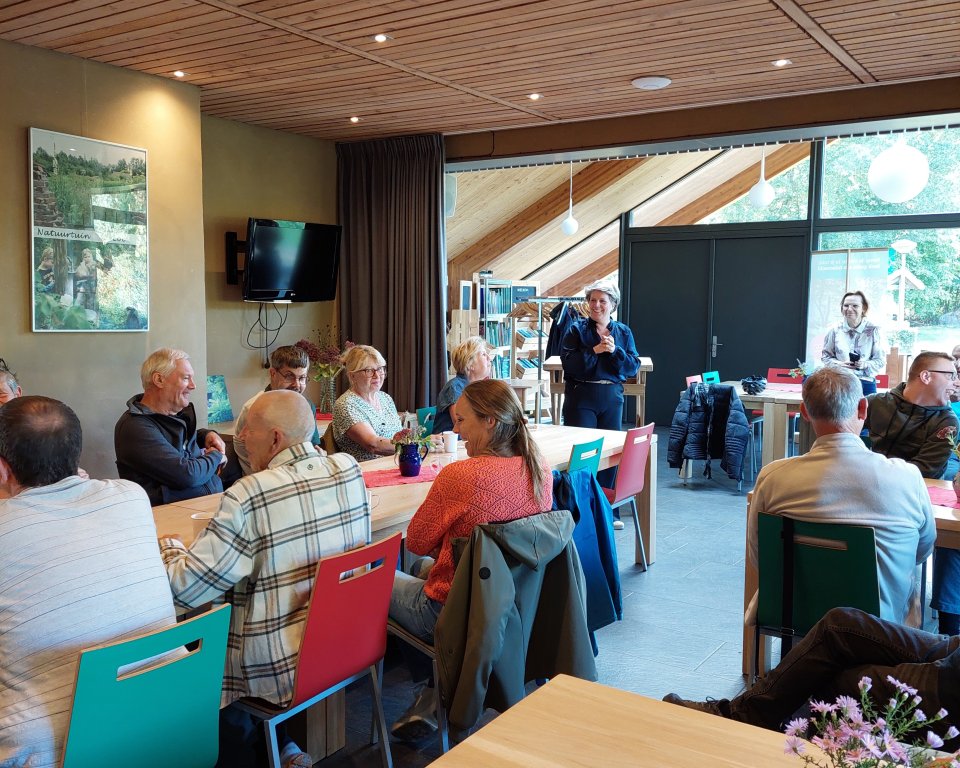 Wethouder Lindy Wijnen staat voor de vrijwilligers die aan tafel zitten bij natuurtuin 't Loo