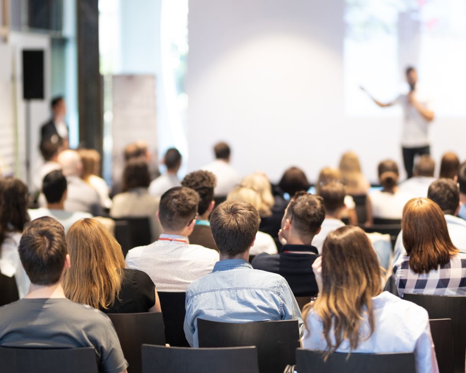 Publiek zit in een zaal te luisteren naar een spreker