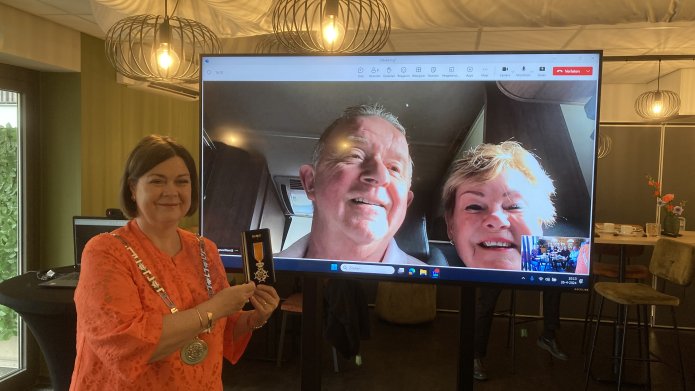 Burgemeester Arinda Callewaert staat met een lintje voor een scherm waarop Hans van den Dungen zichtbaar is