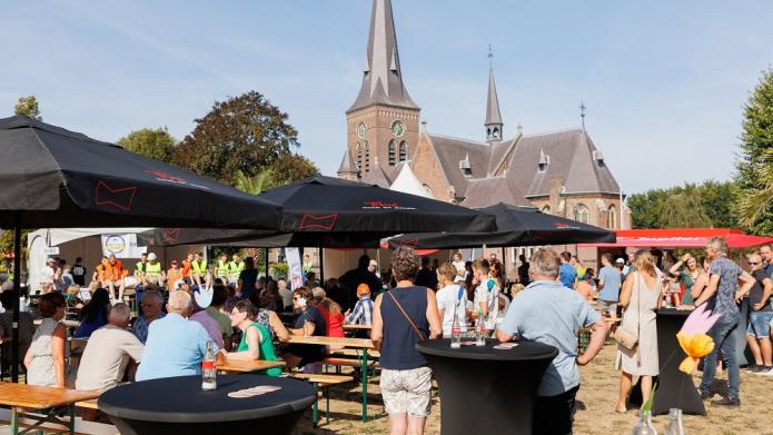 Mensen buiten aan tafels onder parasols met op de achtergrond de kerk van Riethoven