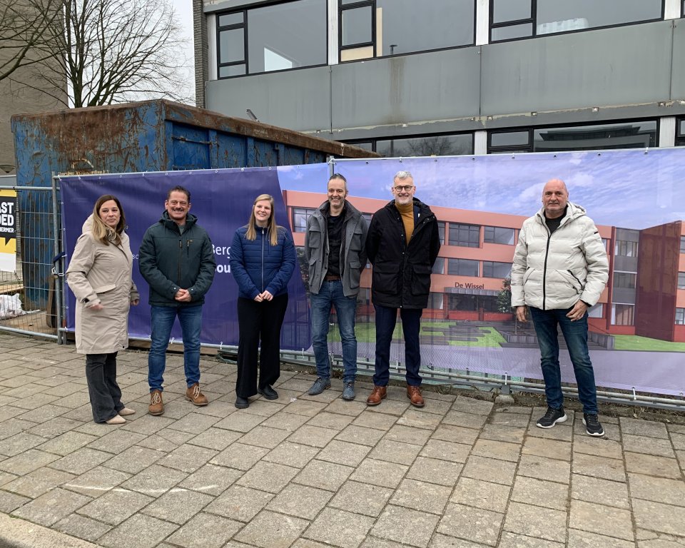 Zes personen op een rij voor een hek met een bouwdoek, daarop staat een afbeelding van een nieuw schoolgebouw. Achter het hek het oude pand en een container.