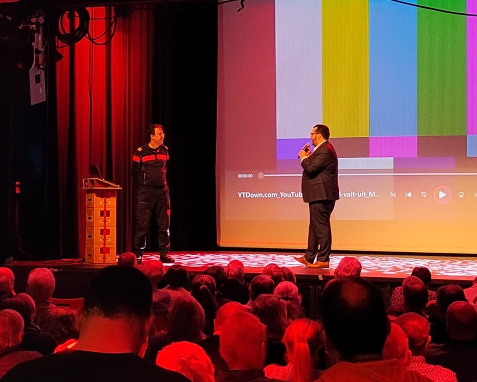 Twee mannen op een podium, op de voorgrond zie je publiek van de achterzijde gefotografeerd.