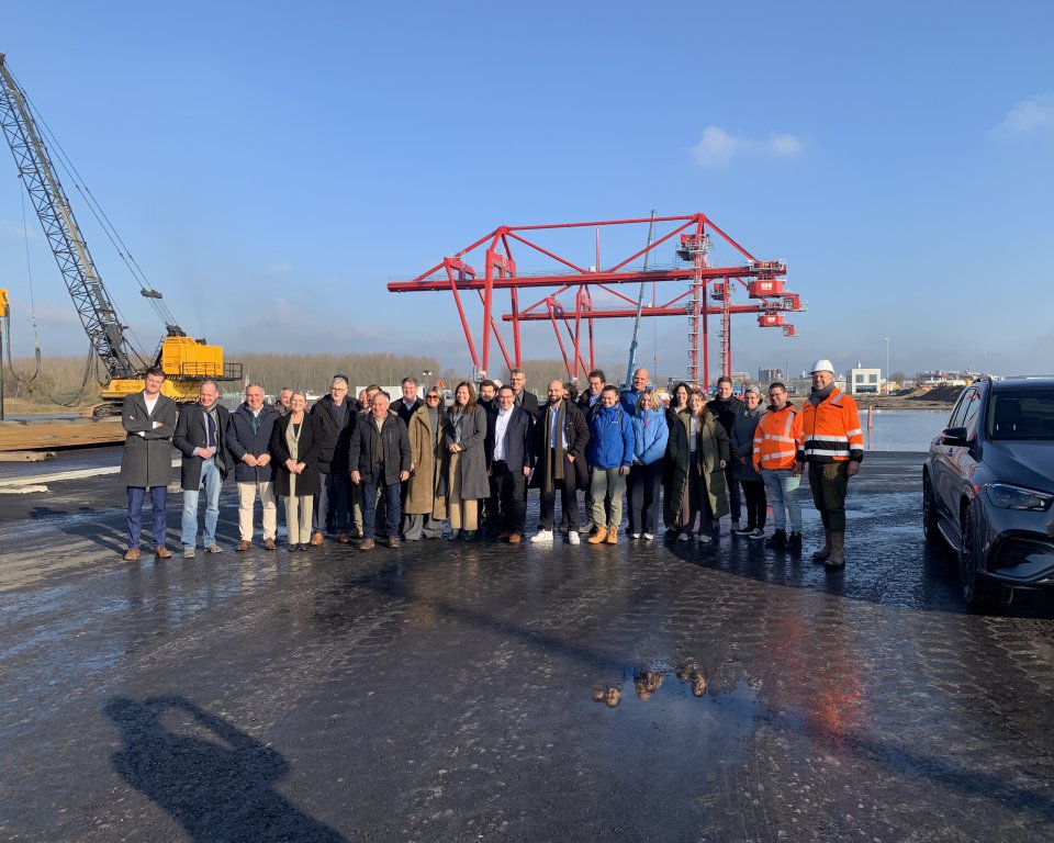 Groep mensen op een bouwplaats, met achter hen een grote gele kraan en in het midden achterin een rode stellage.