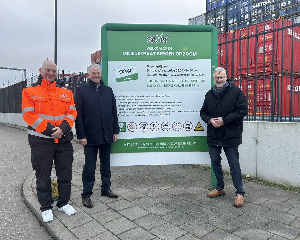 Groot bord in het midden waarop staat 'Milieustraat Bergen op Zoom', daarnaast staan links twee mannen en rechts een man. Op de achtergrond een stapel zeecontainers.