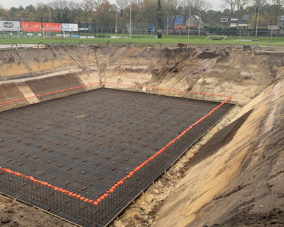 De bouwgrond waar de waterzuiveringsinstallatie gaat komen, diep gat gegraven en op de achtergrond het sportveld.