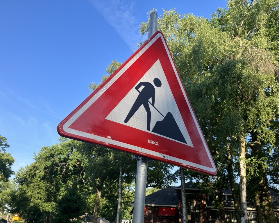 Op de voorgrond een driehoekig wegwerkzaamheden-verkeersbord. Op de achtergrond een rij bomen en huizen.