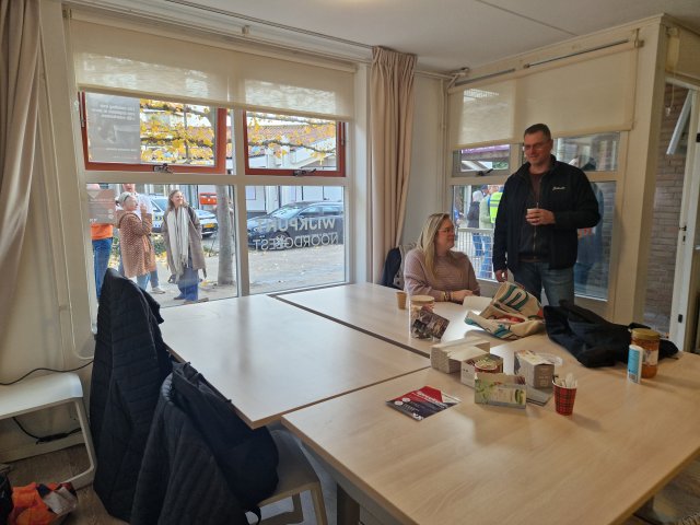 Vier tafels in het wijkpunt, met daaraan 1 zittend en 1 staand persoon. Achter de tafels grote ramen met uitzicht op een deel van de wijk.