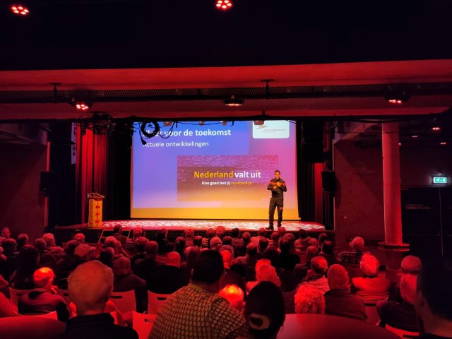 Goedgevulde zaal en op het podium één spreker en op het scherm staat 'Nederland valt uit. Hoe goed ben jij voorbereid?'