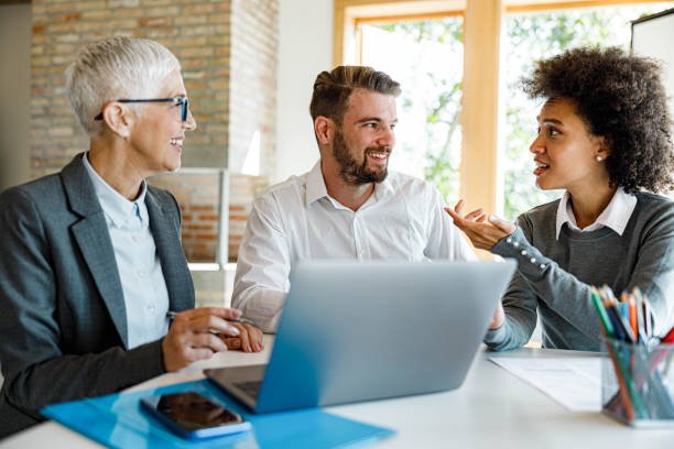 Drie mensen in gesprek aan een tafel, met daarop een map, telefoon, laptop en kleurpotloden.
