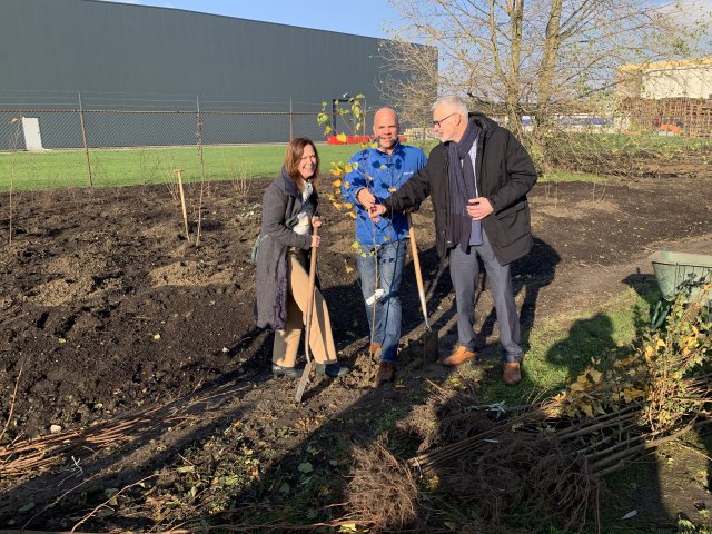 Drie mensen planten een klein boompje, achter hen een groot bedrijfspand.