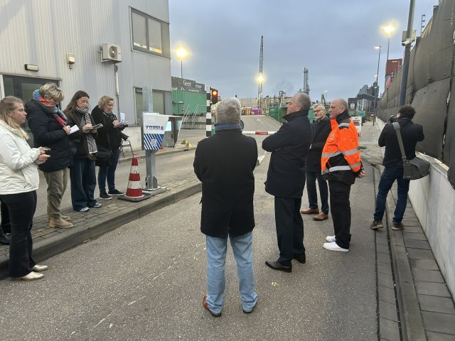 Negen mensen rond de slagboom bij de ingang van de milieustraat, op de achtergrond kranen en containers.