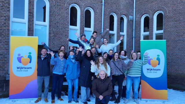 Een groep enthousiaste medewerkers van het wijkteam tussen twee banniers met het logo.