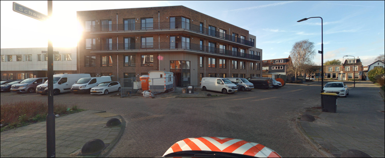 Een T-splitsing met een weg van klinkers. In het midden staat een groot appartementencomplex met geparkeerde auto's ervoor. Rechts zijn in de verte woningen te zien.