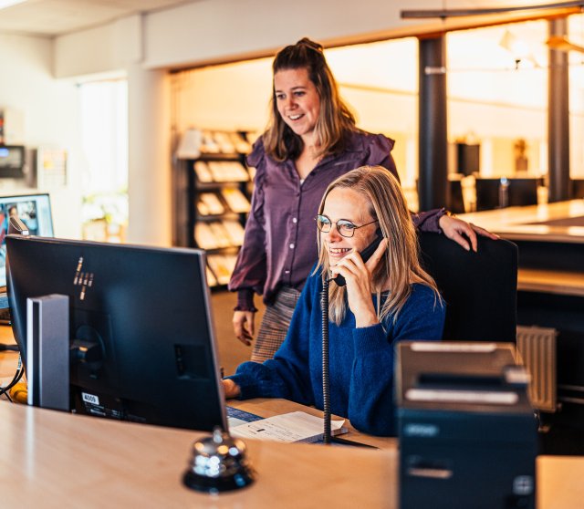 twee vrouwelijke collega's, waarvan één aan de telefoon, kijken naar een computerscherm