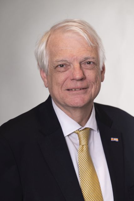 Persoon met donker jasje en gele stropdas poseert voor een neutrale achtergrond