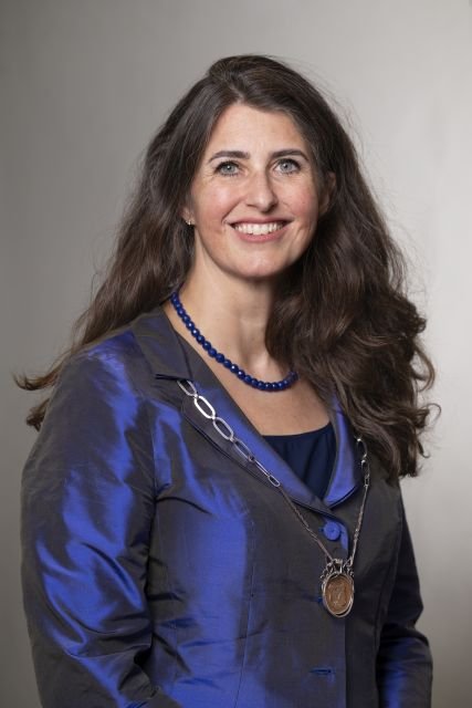 Persoon met ambtsketting en blauwe outfit poseert voor een neutrale achtergrond