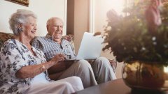 Senioren, man en vrouw, zitten met laptop op schoot op de bank. Ze kijken vrolijk.
