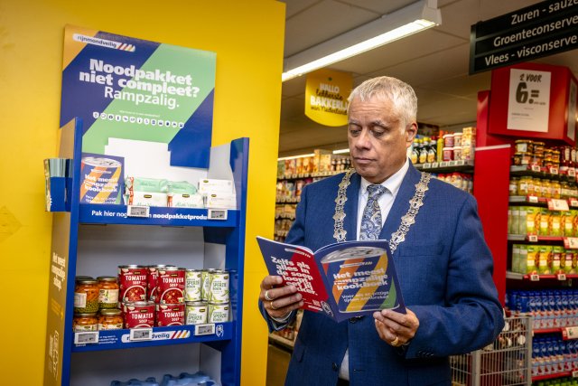 Burgemeester Manusama leest Het meest rampzalige kookboek bij een display voor noodpakketten in een supermarkt