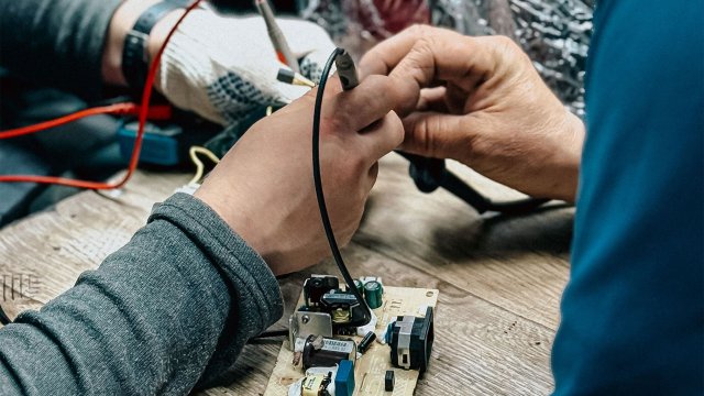 Twee personen repareren een elektronisch onderdeel op een werkbank.