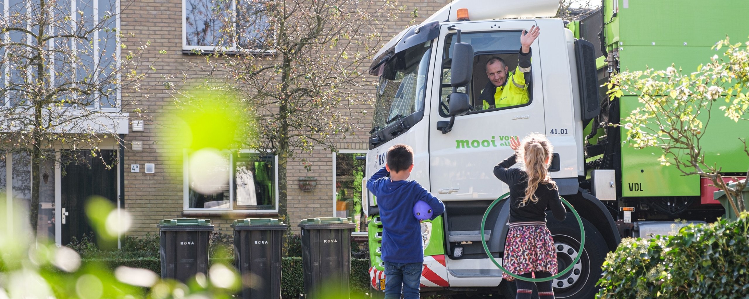 ROVA vuilniswagen rijdt door de straat en chauffeur zwaait naar twee kinderen.