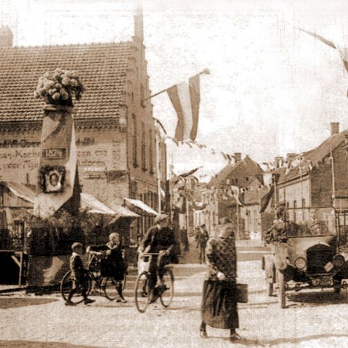 Straatbeeld uit het verleden. Een old-timer, een vrouw die op straat loopt, een fietser en 2 kinderen waarvan 1 met de fiets. Op de achtergrond zijn ook nog een paar wandelende mensen te zien.