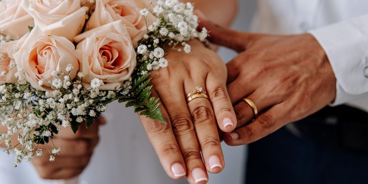 Twee handen van een man en een vrouw met een trouring en de vrouw houd in haar andere had een bosje bloemen vast.