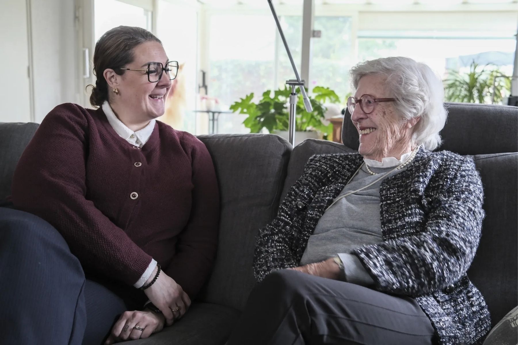 Een mantelzorger in gesprek met een oudere vrouw, samen zitten op een zitbank binnen in een kamer.