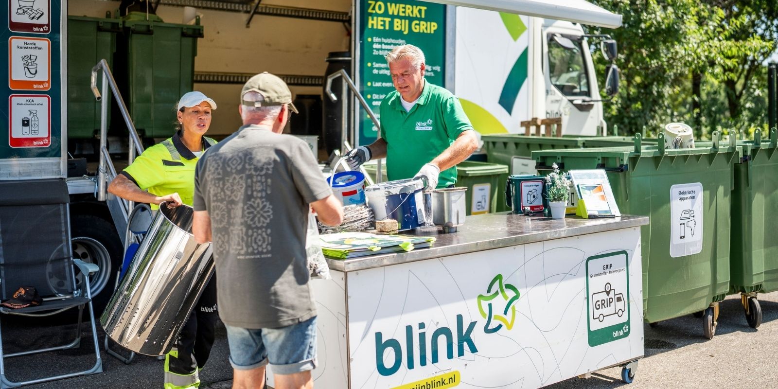 Een persoon levert spullen in bij de GRIP-wagen, waar 2 medewerkers aan het werk zijn en de man helpen.