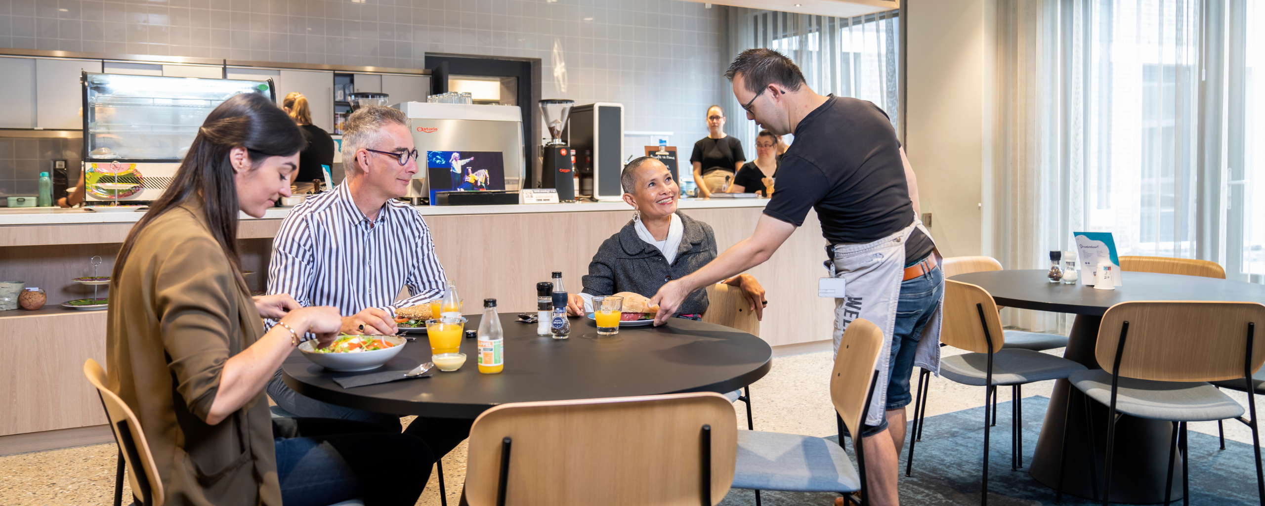 In het lunch- werkcafé zit een aantal collega’s aan tafel te lunchen. De ober serveert een gerecht.