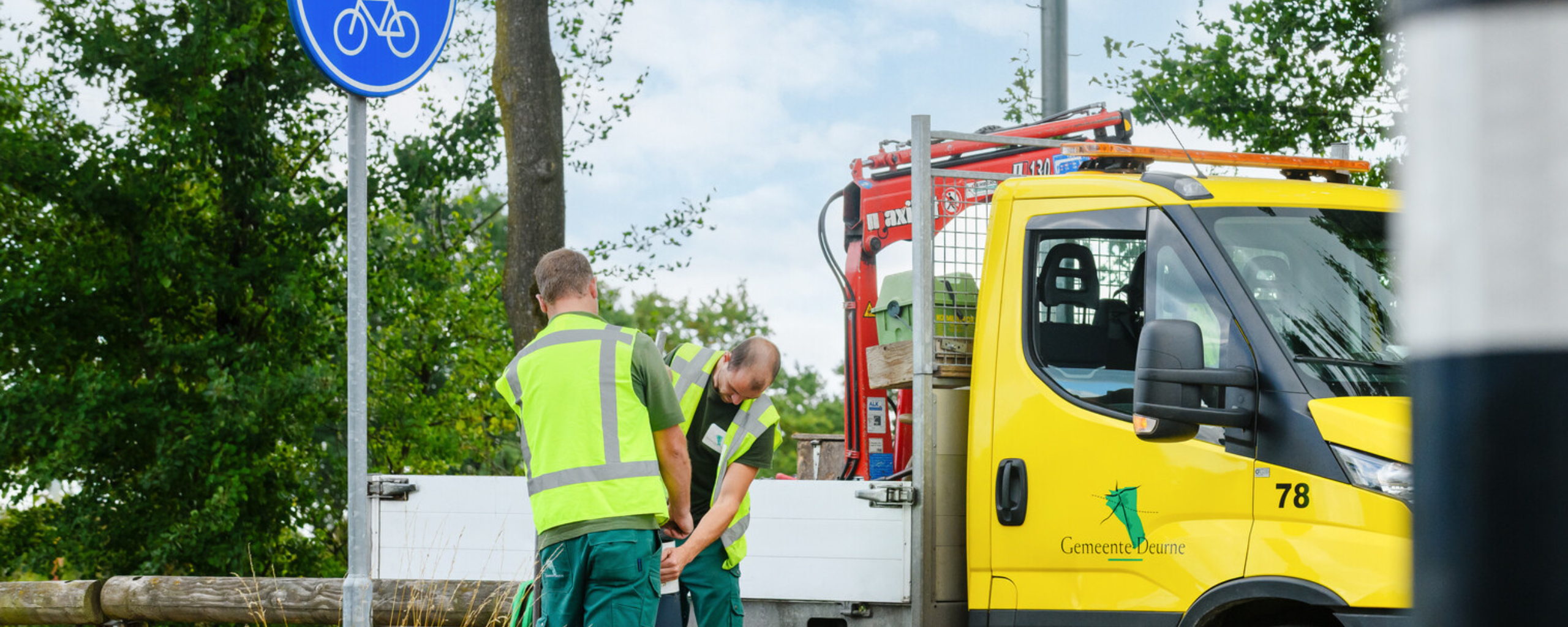 Twee collega's staan voor een dienstauto van gemeente Deurne en tillen een paaltje.
