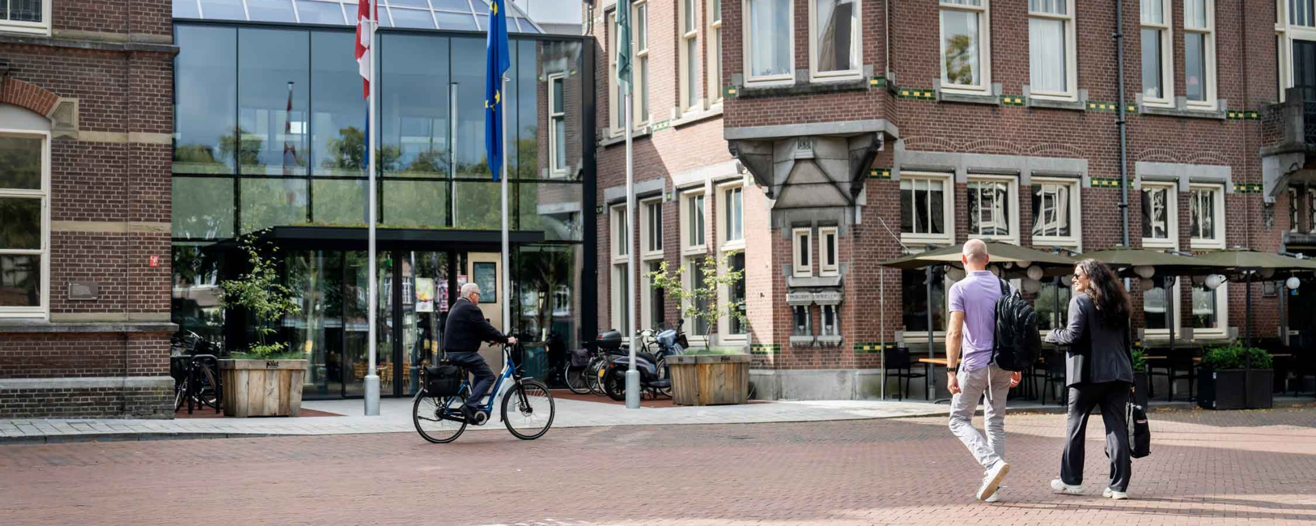 De twee medewerkers zijn onderweg naar de ingang van Het Huis voor de samenleving. Een persoon fietst langs.