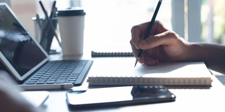 Iemand schrijft iets in een kladblok. Er staat ook een uitgeklapte laptop en er ligt een mobiele telefoon.