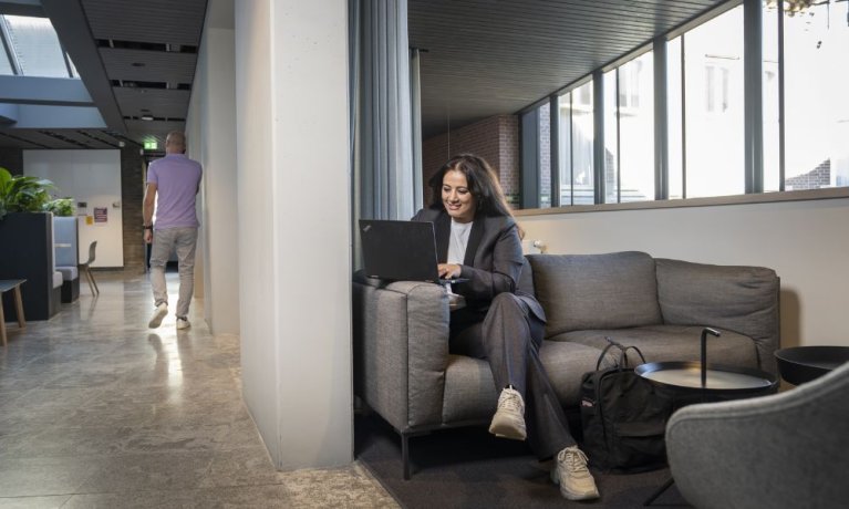 Een medewerkster zit op een bank met haar telefoon, op de achtergrond loopt een andere medewerker.