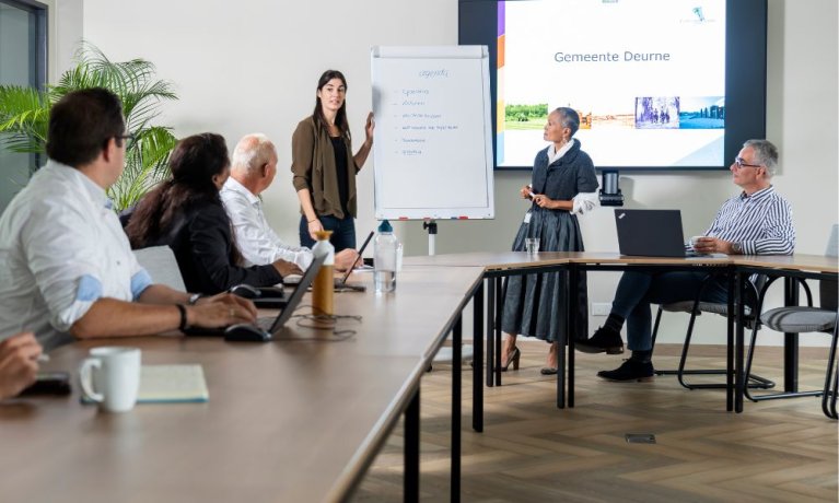 Zes personen in een meeting waarbij 1 persoon een presentatie geeft aan de hand van een whiteboard