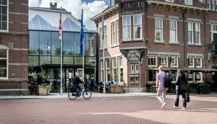 De twee medewerkers zijn onderweg naar de ingang van Het Huis voor de samenleving. Een persoon fietst langs.