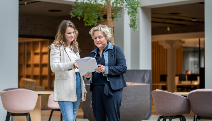 Twee medewerkers staan samen in de gang. Ze kijken en wijzen naar een document die een van de twee vasthoudt.