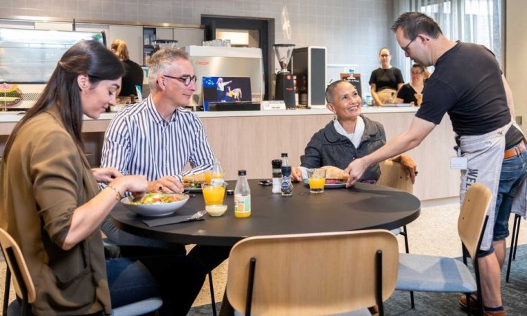 Foto bij de vacature 'medewerker post en archief' gemeente Deurne, foto van een drietal medewerkers in het lunchcafé, met een medewerker van het café die de lunch brengt.