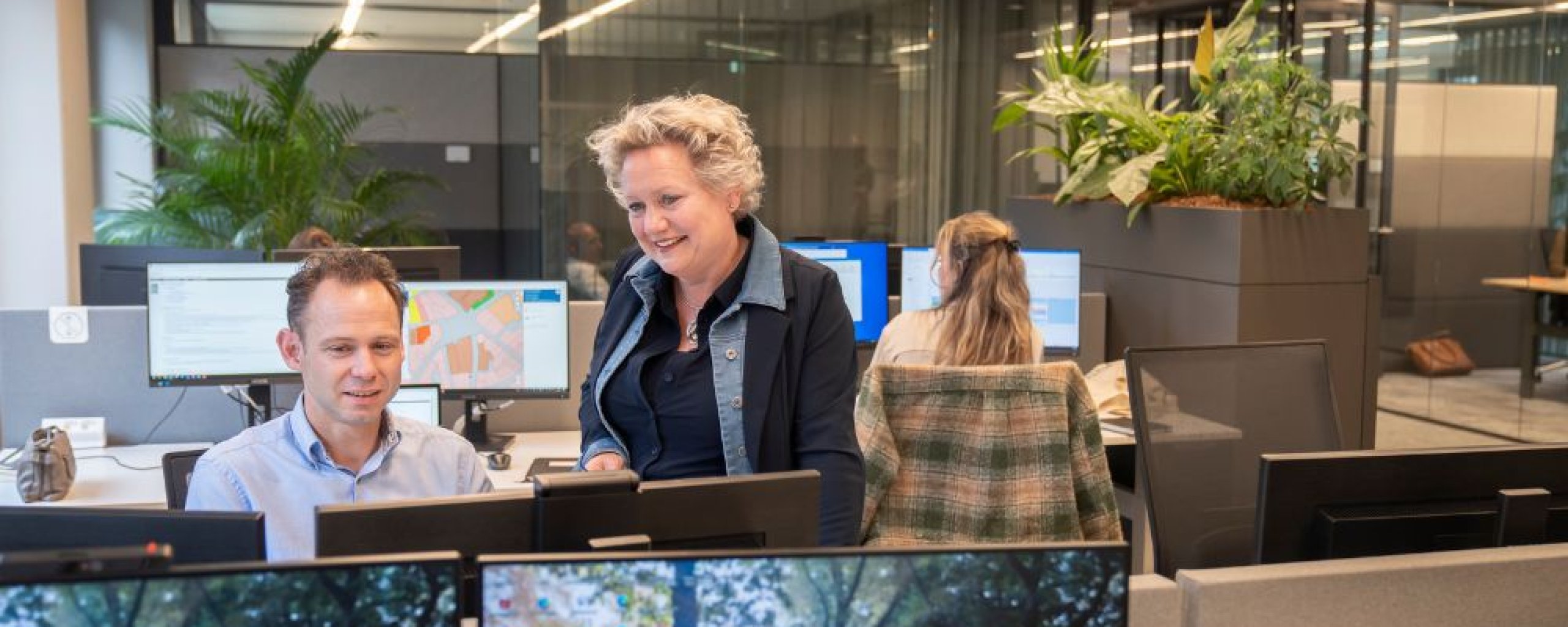 Een medewerker laat trots een andere medewerker wat zien op de computer. De andere medewerker kijkt blij en tevreden toe. Op de achtergrond is een andere medewerker aan het werk 